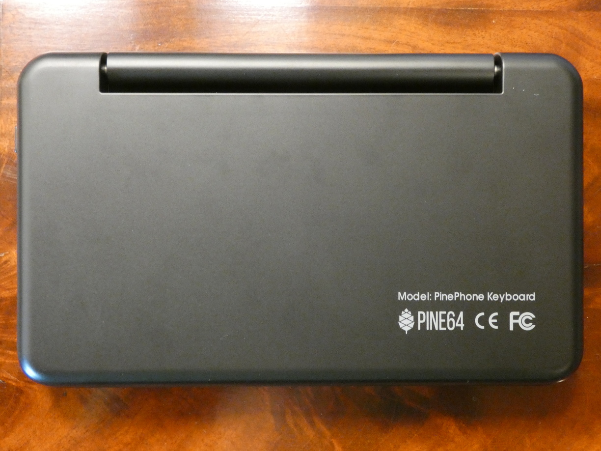 A photo of the empty closed keyboard case from above, now with the bottom side up. This way the long hinge can be seen. In the corner is a medium-sized white print saying 'Model: PinePhone Keyboard', the Pine64 logo and CE and FC logos.